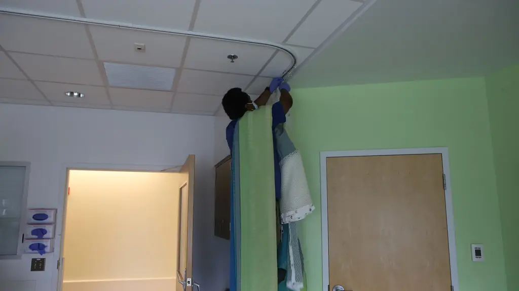 Installer at the ceiling track re-hanging a privacy curtain in a green-walled patient room